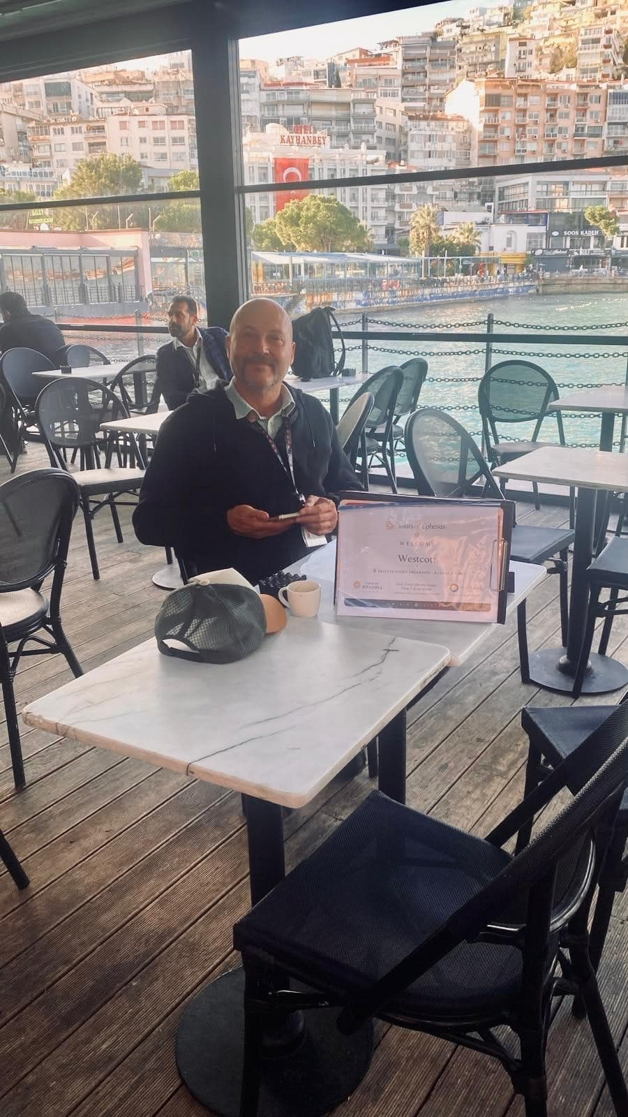 Licensed guide Serdar Atalan waiting on the Starbucks terrace at Kuşadası cruise port with a Tours of Ephesus welcome sign for his two Azamara guests, before a private Ephesus tour, 29 October 2026