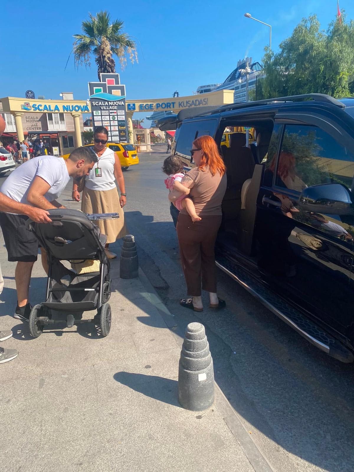 A family with a baby and a stroller boarding a black Mercedes V-Class at the Ege Port Kuşadası exit with their licensed guide Fulya Tamer, a cruise ship behind, at the start of a private Ephesus tour, 26 August 2026