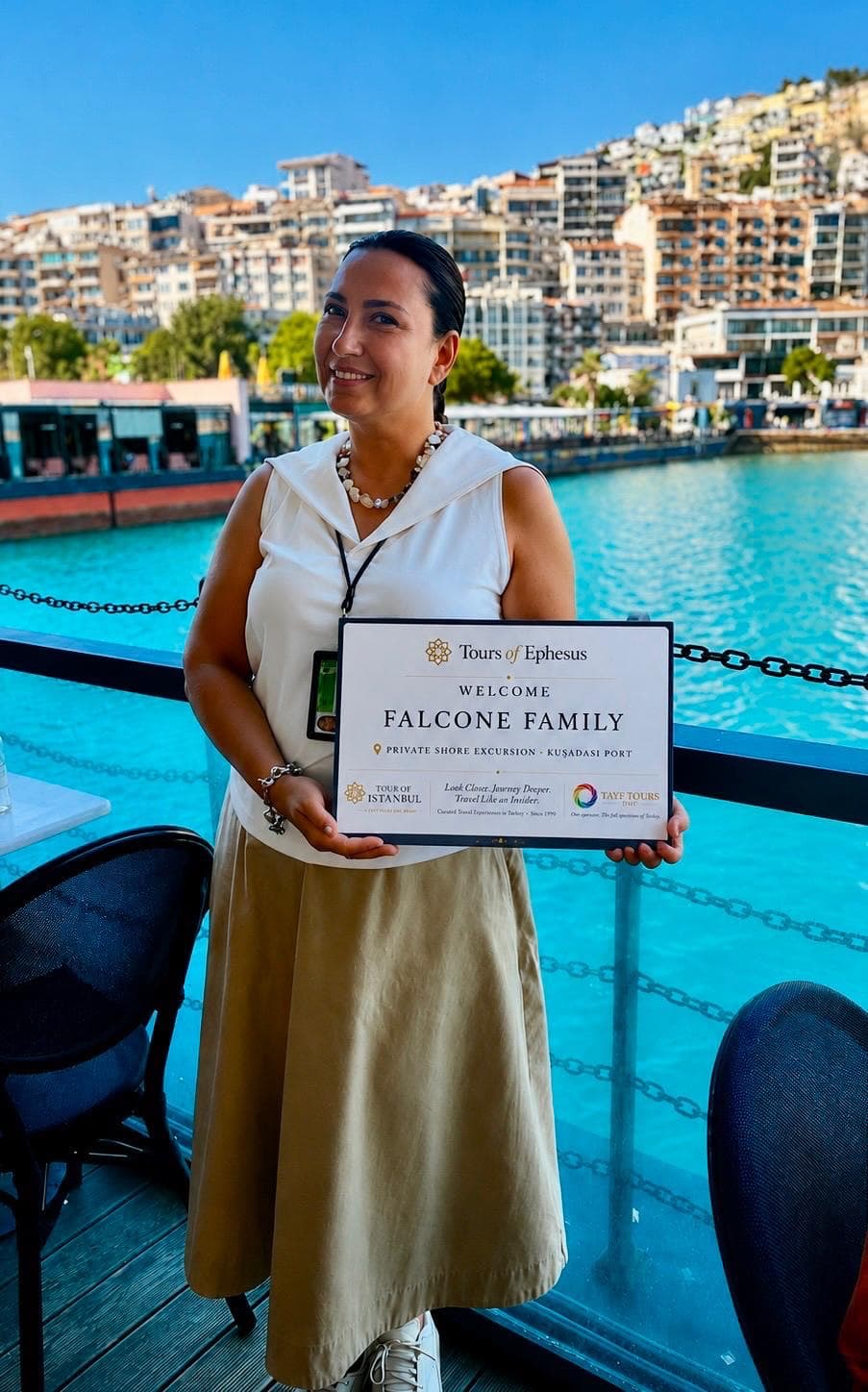 Licensed guide Fulya Tamer holding a Tours of Ephesus welcome sign for a private shore excursion at Kuşadası port, the harbour and the town behind her, 26 August 2026