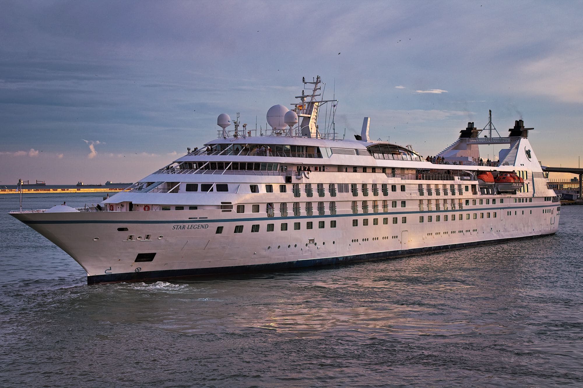 Windstar cruise ship Star Legend, white with a teal stripe and her name on the bow, leaving the port of Barcelona in the early evening
