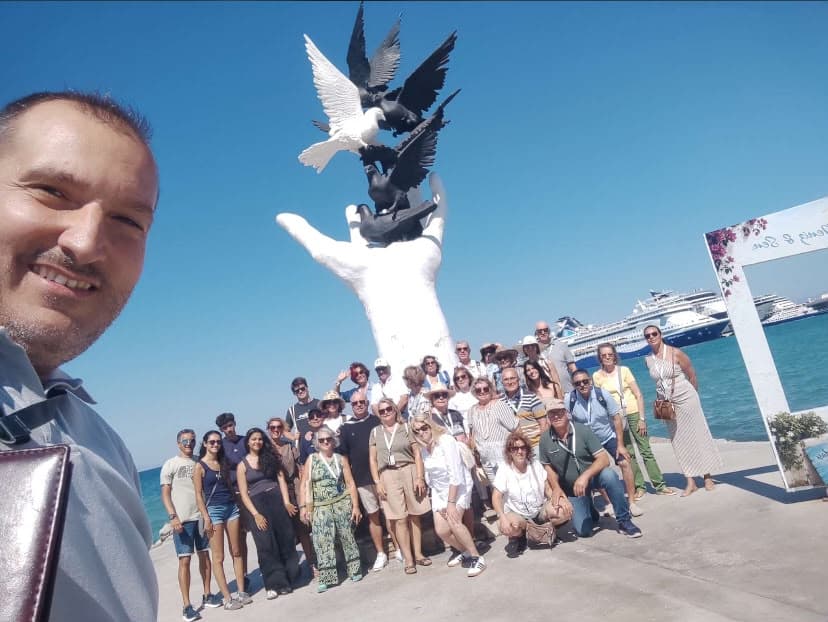 Our guide Gökhan Özden with the whole tour group at the dove sculpture on the Kuşadası harbour front, cruise ships docked behind, July 2026