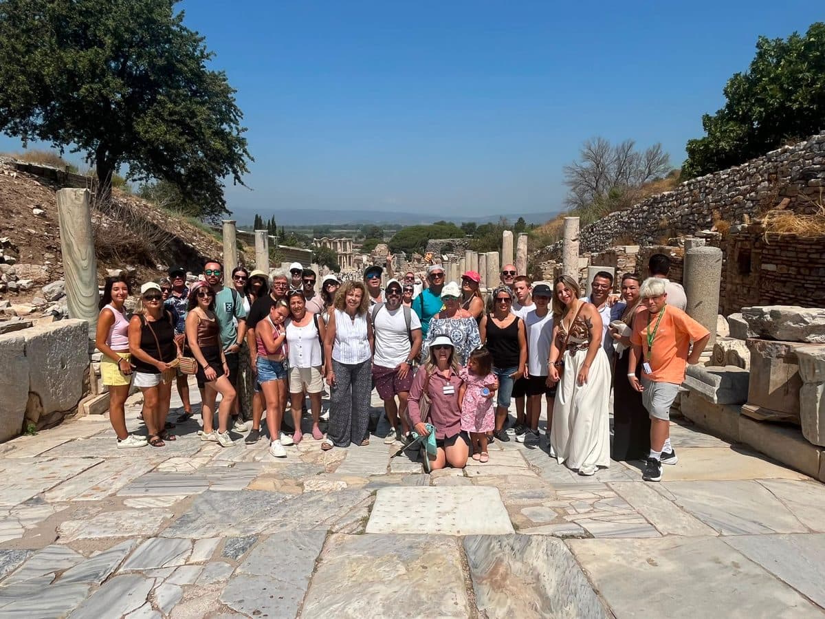 Our Spanish-speaking licensed guide Ebru Özkan kneeling front and centre with a large group of MSC Divina guests on the marble paving of Curetes Street in Ephesus, the Library of Celsus far below, 3 August 2026