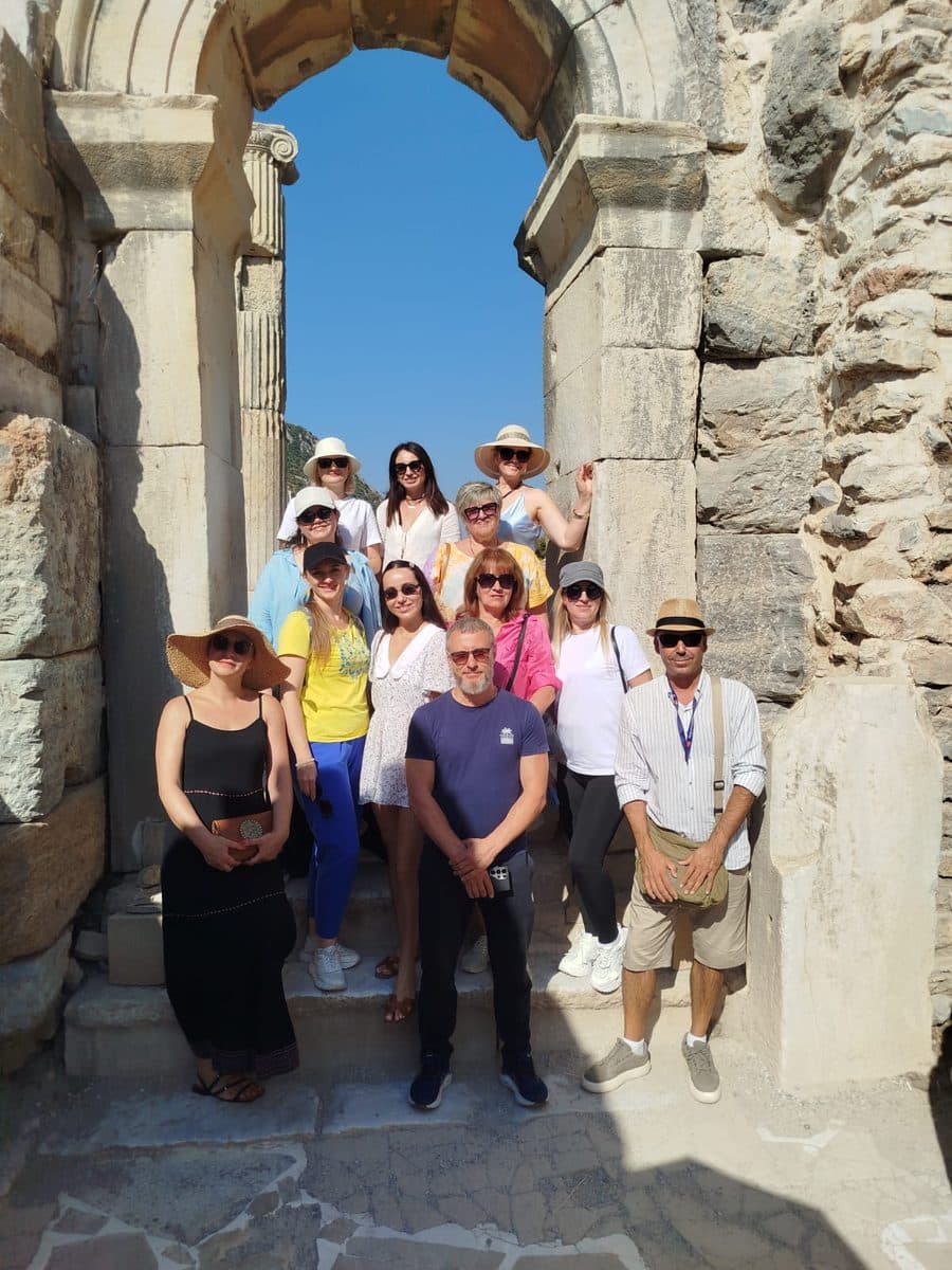 Russian-speaking licensed guide Cihan Yaşkaya, on the right in a striped shirt and straw hat, with eleven MSC Divina guests under a Roman stone archway at Ephesus, on a private Ephesus, House of the Virgin Mary and Temple of Artemis tour in Russian from Kuşadası cruise port, 21 September 2026