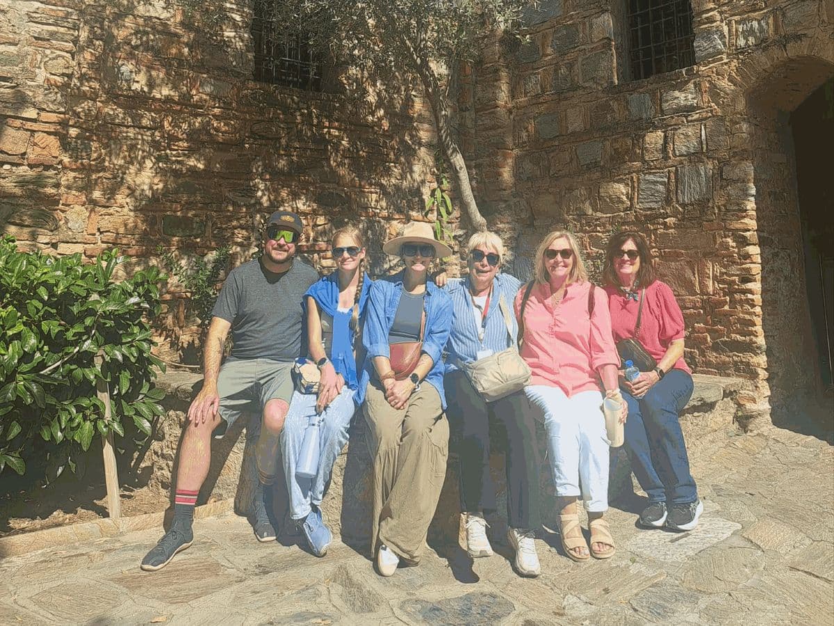 Licensed English-speaking guide Burçak Akdemir, fourth from left in a striped blue shirt, seated with five Odyssey of the Seas guests beside the stone walls of the House of the Virgin Mary, on a private Ephesus, House of the Virgin Mary and Temple of Artemis tour from Kuşadası cruise port, 3 October 2026