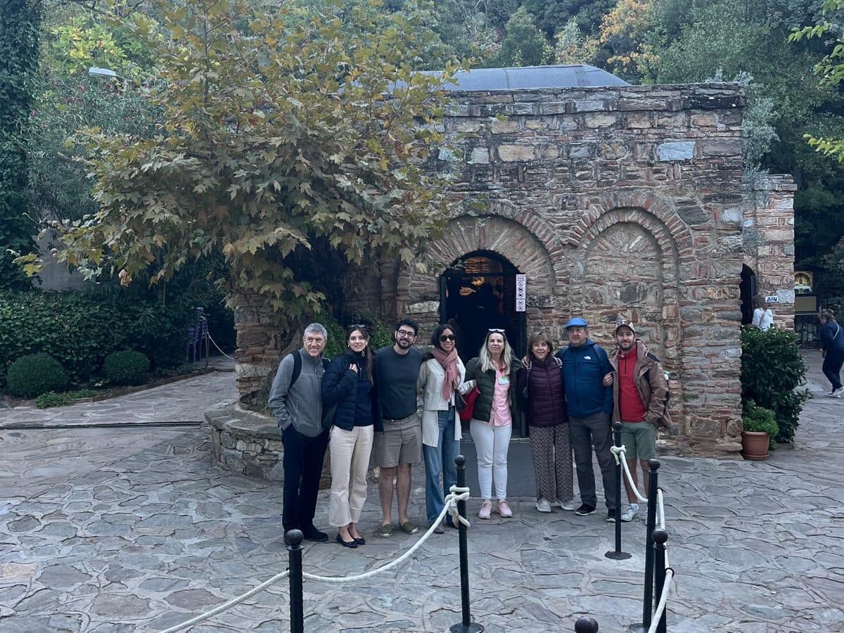 Spanish-speaking licensed guide Ebru Özkan with seven Holland America Oosterdam guests from Andorra, Ebru in the centre, in front of the stone chapel of the House of the Virgin Mary on Mount Koressos, on a private tour in Spanish from Kuşadası cruise port, 20 October 2026