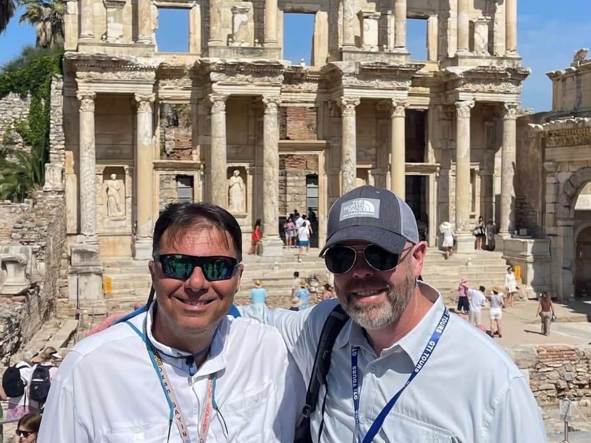 Licensed English-speaking Ephesus tour guide Ozan Sayın, on the left in sunglasses and a white shirt, with his arm around a guest on the plaza in front of the Library of Celsus at Ephesus, the Gate of Mazaeus and Mithridates at the right