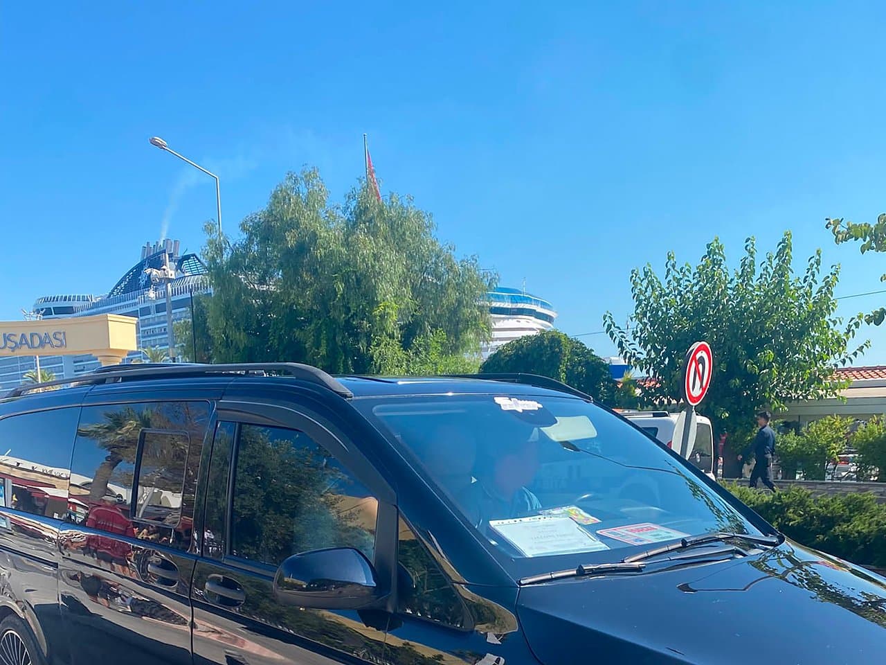 Black tour van waiting at the Kuşadası cruise port exit with a family name sign in the windscreen and a cruise ship behind