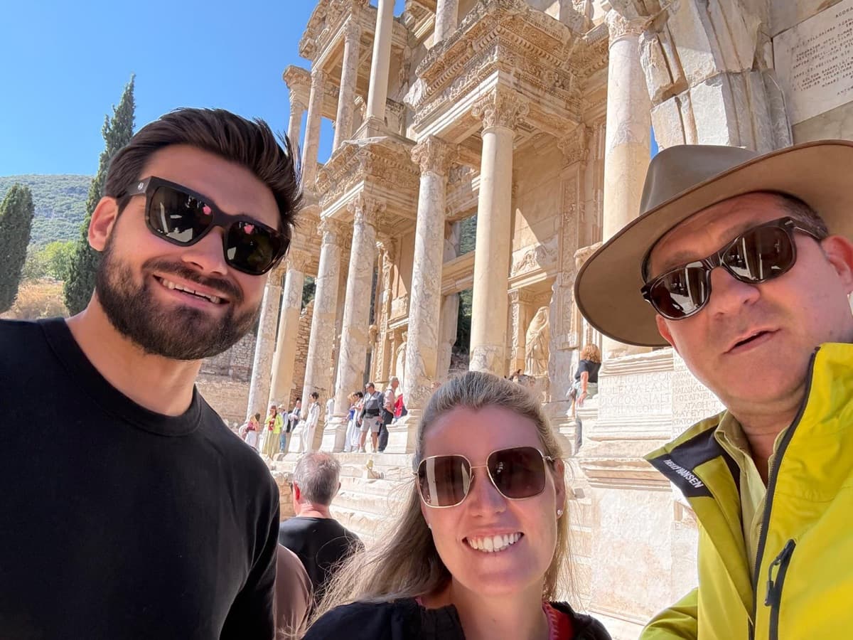 Licensed English- and Russian-speaking guide Mehmet Tunalı in a selfie with two Ritz-Carlton Evrima guests in front of the Library of Celsus, on a private Ephesus, House of the Virgin Mary, Temple of Artemis and traditional Turkish lunch tour with a carpet-weaving demonstration from Kuşadası cruise port, 2 October 2026
