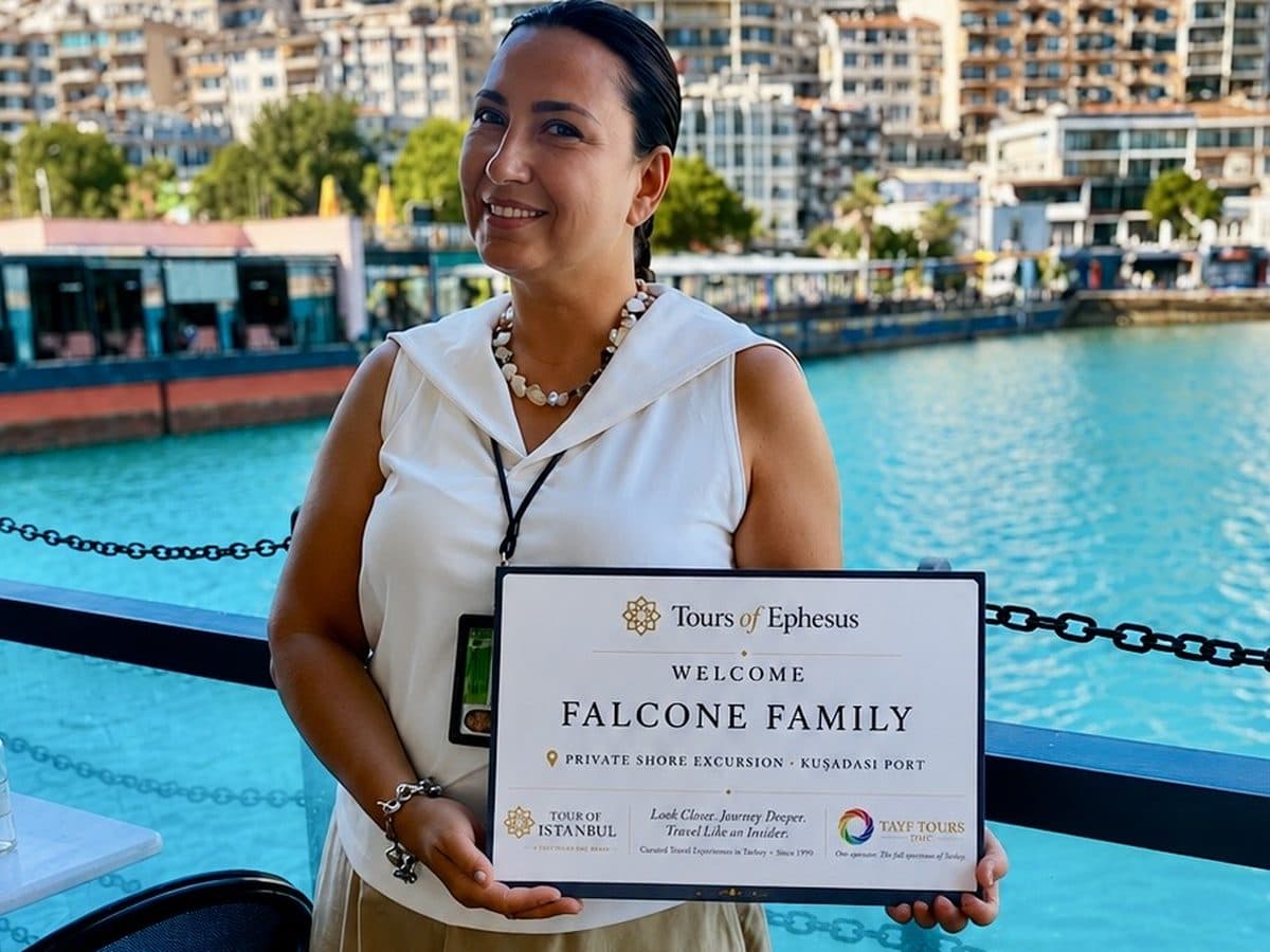 Tours of Ephesus guide Fulya Tamer holding a printed welcome board with the guests' name on the Starbucks terrace at Kuşadası cruise port, the marina behind her