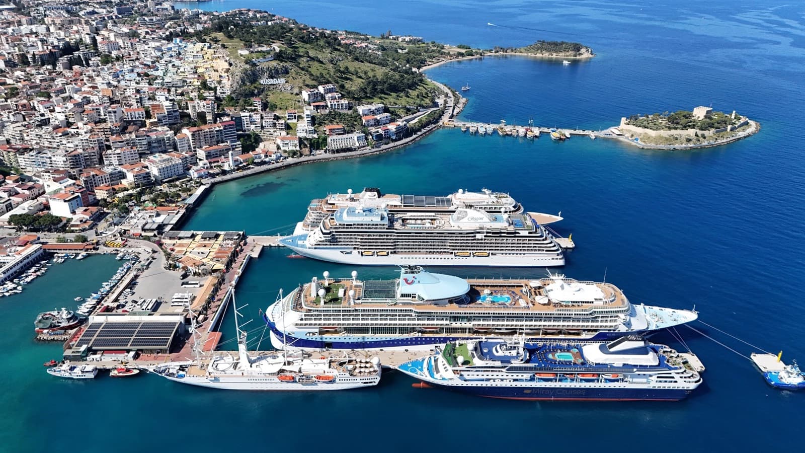 Aerial view of Kuşadası cruise port with several cruise ships docked, Pigeon Island and the Kuşadası hillside beyond