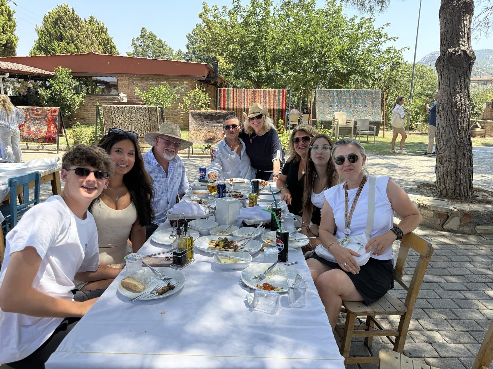 Family sharing a long Turkish lunch table in a shaded garden near Ephesus, handwoven carpets drying behind
