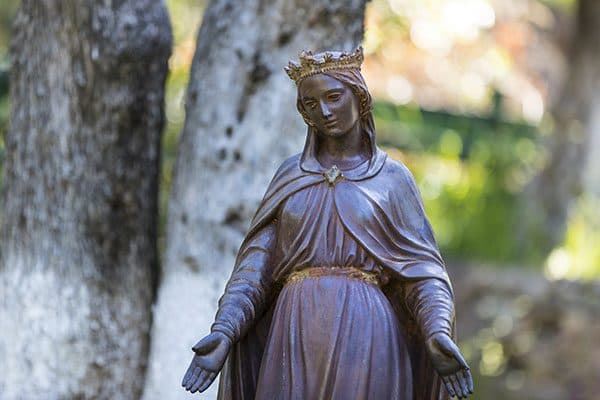Crowned bronze statue of the Virgin Mary in the shrine garden, olive trunks soft in the background