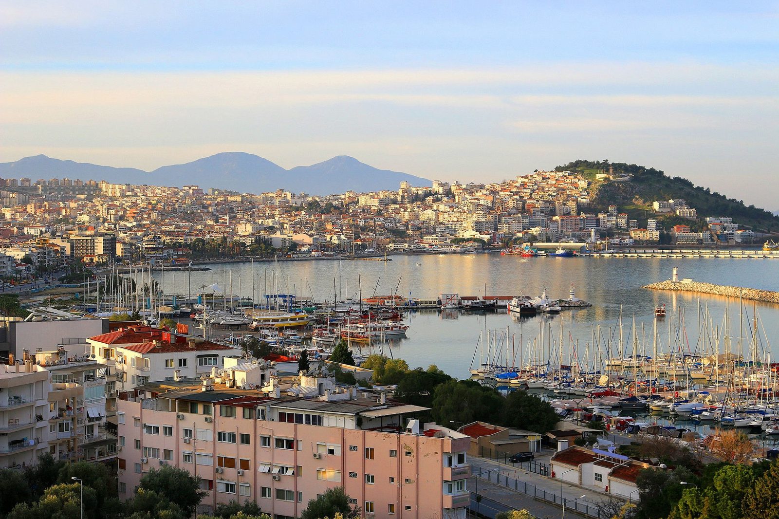 Kuşadası bay at golden hour: the marina full of sailboats, the breakwater lighthouse, and hillside neighbourhoods rising in tiers behind the harbour